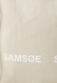 Béžová plátenná taška s textom "SAMSNØE" v tučnom bielom písmene. Materiál má textúrovanú väzbu s jednoduchým dizajnom.