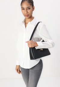 Black faux leather handbag with a triangular shape and a gold clasp, worn with a white button-up shirt and gray jeans.