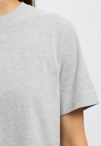 Gros plan d’une personne portant un T-shirt uni gris clair à manches courtes avec un col rond sur un fond uni.