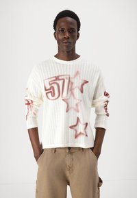 Maglione bianco di grandi dimensioni con motivo a stelle e grafica "57" in rosso. Tessuto a maglia testurizzato con maniche lunghe. Indossato con pantaloni cargo beige.