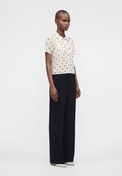 Femme portant un chemisier blanc à manches courtes avec des pois noirs, un pantalon noir large et des chaussures blanches pointues, debout devant un fond uni.