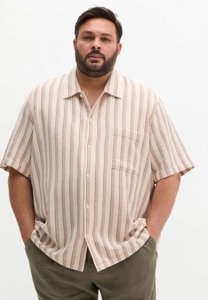 Mężczyzna z brodą w beżowej koszuli z krótkim rękawem z czerwonymi pionowymi paskami i oliwkowozielonych spodniach, stojący z rękami w kieszeniach.