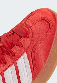 Κόκκινο αθλητικό παπούτσι με υφασμάτινο επάνω μέρος με υφή, λείες κορδόνια, τρεις λευκές ρίγες και σόλα από καουτσούκ χρώματος καραμέλας.