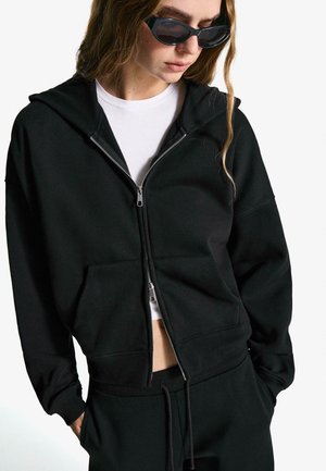 Zwarte rits-hoodie van een katoenmengsel, met een voorkantzak en ribgebreide manchetten. Draag ik over een wit shirt, gecombineerd met zwarte broek.
