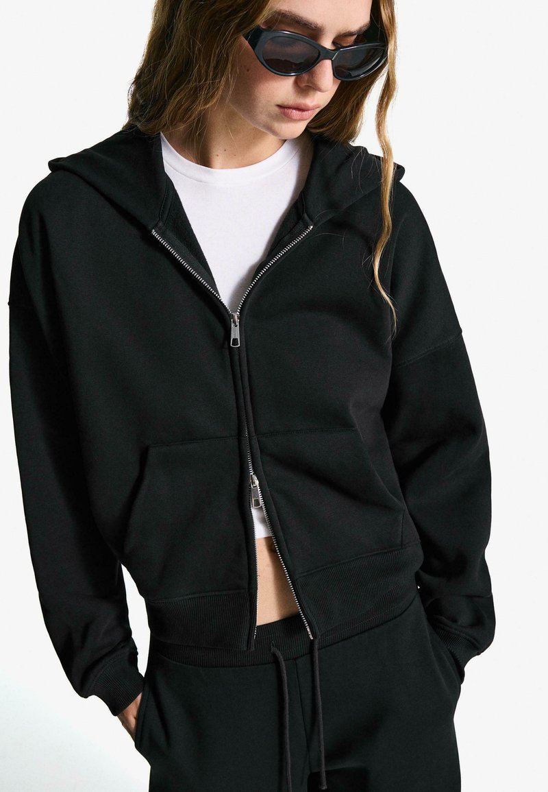 Black zip-up hoodie made of cotton blend, featuring a front pocket and ribbed cuffs. Worn over a white shirt, paired with black pants.