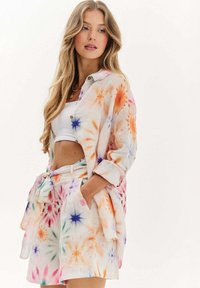 Žena oblečená v bielom crop top a farebnej košeli s uzlom v štýle tie-dye, spolu s rovnakými krátkymi nohavicami s vreckami, stojí na jednofarebnom pozadí.