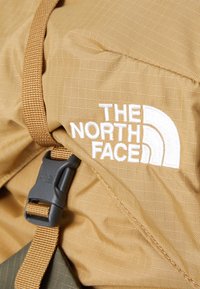 Light brown hiking backpack with textured nylon fabric, featuring a black plastic buckle and white embroidered logo detail.