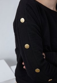 Maglione nero in tessuto testurizzato con bottoni decorativi dorati sulle maniche. Il design presenta una vestibilità rilassata e dettagli a coste.