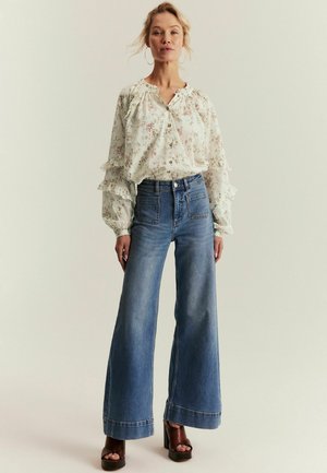 Kvinde iført en langærmet bluse med blomsterprint og flæsedetaljer, højtaljede viddebukser og brune plateauhøjhælede sko, stående mod en lys baggrund.