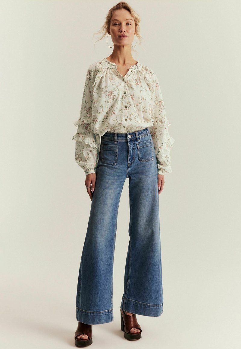 Nainen, joka pukeutunut kukalliseen pitkähihaiseen puseroon, jossa on röyhelötietoja, korkeavyötäröisiin leveälahkeisiin farkkuihin ja ruskeisiin korollisiin platinalehtoihin, seisoo vaaleaa taustaa vasten.