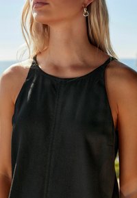 Haut noir sans manches avec un col rond, présentant un léger éclat et un détail de couture verticale, associé à des boucles d'oreilles créoles.