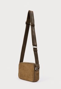 Καφέ σουέντ τσάντα ώμου με ορθογώνιο σχήμα, υφή επιφάνειας και ρυθμιζόμενο λουράκι με μεταλλική πόρπη. Κλείνει με φερμουάρ στην κορυφή.