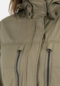 Gros plan sur une veste vert kaki avec des zips noirs verticaux et un col à boutons-pression, portée par une personne aux cheveux bruns.