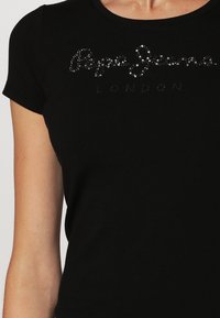 Mustat istuvat lyhythihaiset paidat, joissa on kimaltelevat strassitekstit "Pepe Jeans London" rinnassa, päällä henkilöllä.