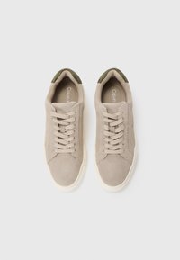 Beige suededsneakers med olivengrønne accenter ved kraven, med flade snørebånd, en tekstureret overdel og en cremet gummisål.