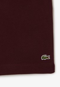 Bordeaux katoenen T-shirt met een klein geborduurd alligatorlogo in groen en rood, aan de onderzoom, met een gladde textuur.