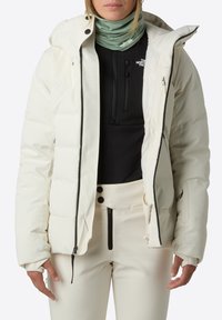 Biała izolowana kurtka z kapturem, zapięcie na zamek i bocznymi kieszeniami. Czarna warstwa bazowa i jasnoszara osłona na szyję noszone pod spodem.