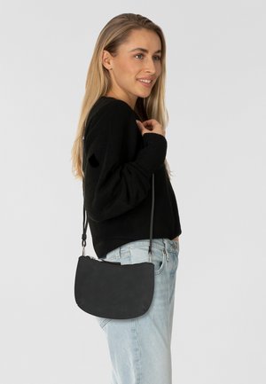 Sac à épaule en cuir noir au design courbé, avec fermeture éclair et bandoulière réglable, posé devant un fond de jean clair et un pull noir.