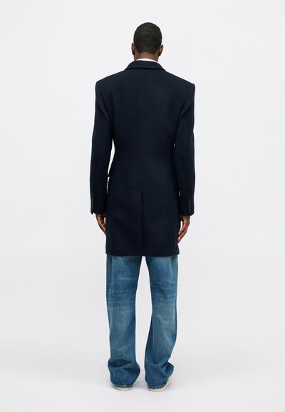 Navy wool overcoat with a long design, notched collar, and two-button sleeves; paired with light blue wide-leg jeans.