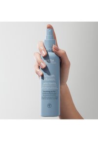 Ljusblå sprayflaska som hålls i hand, märkt "Aveda Smooth Infusion," med detaljer i vit text, cylindrisk form och spraymunstycke.