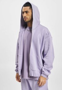 Sweat à capuche zippé violet clair avec un design spacieux, une texture douce, de grandes poches avant et une capuche confortable. Porté sur un t-shirt assorti.