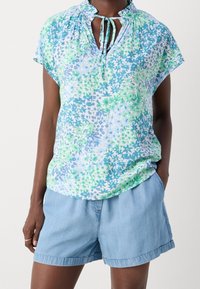 Blouse à fleurs bleues et vertes avec un col froncé et un détail de lien, fabriquée en tissu léger, manches courtes en forme de cap, et coupe ample.