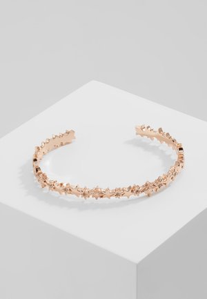 Bracelet jonc ouvert en or rose avec des motifs étoilés et floraux texturés, présenté sur un piédestal géométrique blanc sur fond gris clair.