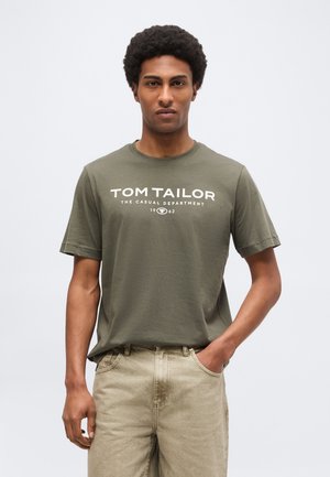 Άνδρας που στέκεται αντικριστά φορώντας μια ελιά πράσινη μπλούζα «Tom Tailor» και μπεζ παντελόνι με το ένα χέρι στην τσέπη.