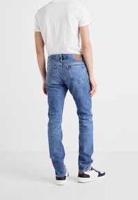 Person i ljusblå Lee jeans, vit t-shirt och vita sneakers med svarta detaljer, stående mot en enkel vit bakgrund.