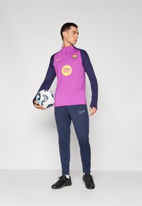 T-shirt desportiva de mangas compridas roxa com mangas azul-marinho e um logotipo do Barcelona, emparelhada com calças de treino Nike azul-marinho e sapatos de futebol pretos. A segurar uma bola de futebol.