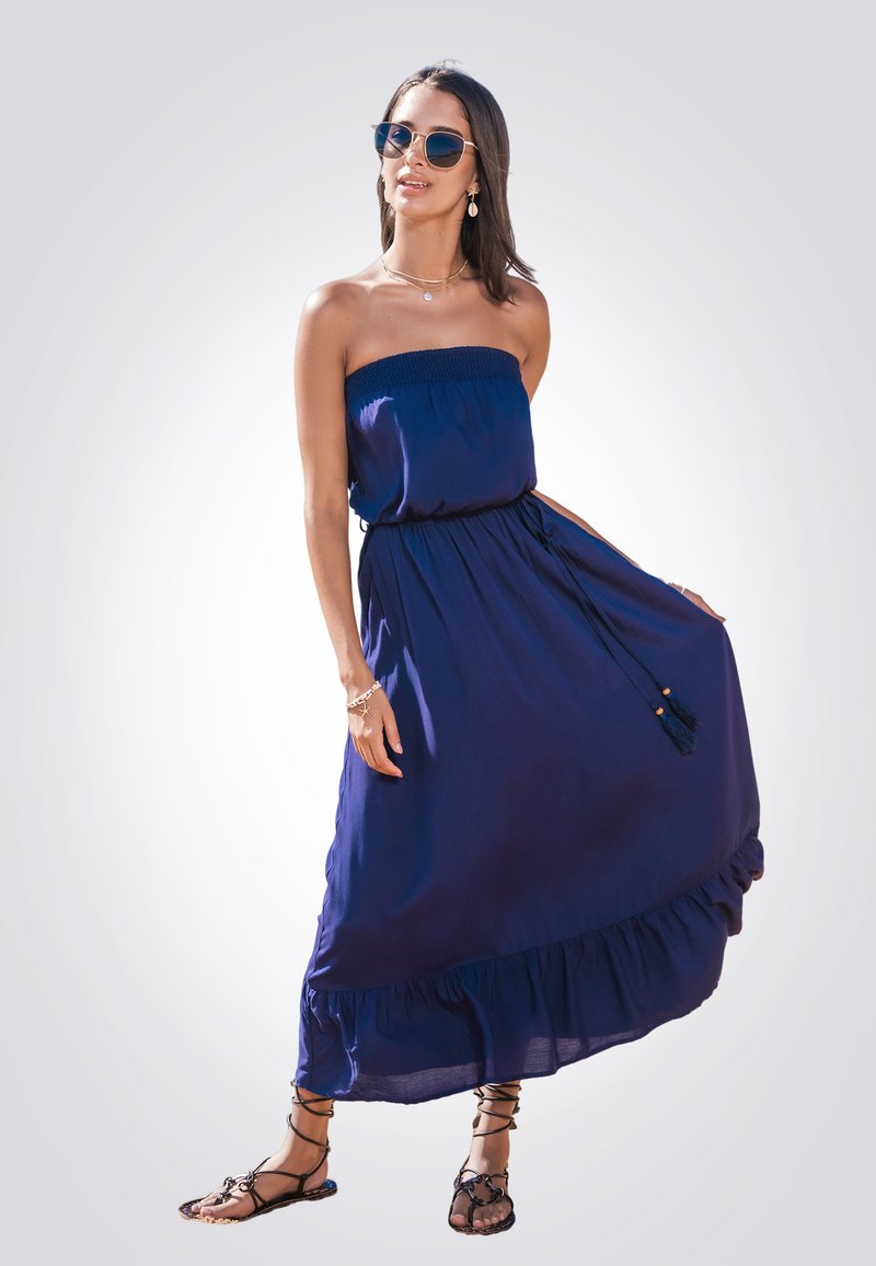 Abito lungo senza spalline di colore blu navy con corpetto arricciato, vita regolabile con cordino e orlo a volants. Abbinato a sandali neri con cinturini e occhiali da sole.