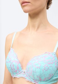 Κοντινό πλάνο γυναίκας που φοράει ανοιχτό μπλε σουτιέν από δαντέλα με ροζ λουλουδάτα σχέδια και λεπτές τιράντες στους ώμους, σε ουδέτερο φόντο.