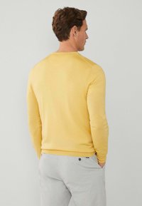 Suéter amarillo con una textura suave y mangas largas, visto desde la parte posterior. Combinado con pantalones gris claro. Cuello redondo simple.