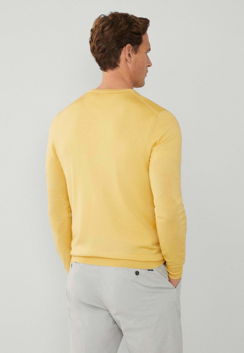 Suéter amarillo con una textura suave y mangas largas, visto desde la parte posterior. Combinado con pantalones gris claro. Cuello redondo simple.