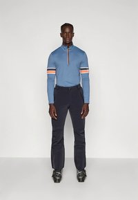 Plava majica s dugim rukavima i zatvaračem, s narančastim i bijelim prugama na rukavima, uparena s crnim hlačama i skijaškim čizmama, stoji na jednobojnoj pozadini.