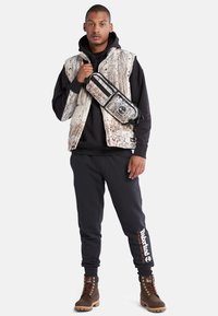 Gepolsterte Weste mit rindähnlichem Muster, schwarzer Kapuzenpullover, dunkle Jogginghose und braune Schnürstiefel. Umhängetasche mit Logodetail.