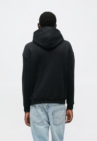 Felpa nera con cappuccio, tasca marsupio frontale, maniche larghe e coulisse, abbinata a jeans di denim azzurro chiaro.