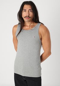 Tricou din bumbac gri, fără mâneci, cu decolteu rotunjit, având un mic logo roșu și albastru pe pieptul stâng. Modelul poartă blugi negri.