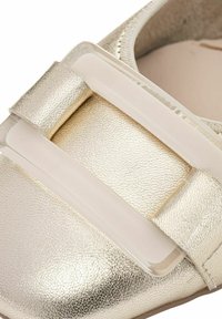 Zapato plano metálico dorado con una parte superior de cuero texturizado, que presenta una correa de acento de plástico transparente y un diseño de puntera redondeada.