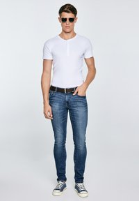 Junger Mann mit Sonnenbrille, weißem kurzärmligen Hemd, blauer Jeans mit Gürtel und blauen Turnschuhen, der vor einem schlichten hellen Hintergrund steht.