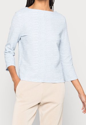 Frau trägt einen hellblauen, strukturierten Langarmtop und beige Hose, mit einer Hand in der Tasche, stehend vor einfarbigem Hintergrund.