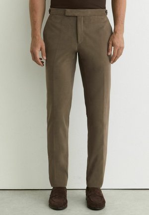 Mann trägt schlanke braune Hose und dunkelbraune Schuhe, steht vor einer schlichten weißen Wand.