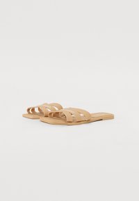 Tan slide-mokkasiinit, joissa on kolme pystysuoraa hihnaa ja litteä pohja, valmistettu teksturoidusta materiaalista. Ylähihnassa on hienovarainen brändäys.