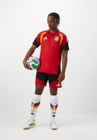 Κόκκινη και μαύρη αθλητική στολή ποδοσφαίρου με κίτρινες λεπτομέρειες, που περιλαμβάνει δικτυωτή φανέλα, αντίστοιχα σόρτς, λευκές κάλτσες με σχέδιο και παπούτσια με καρφιά.