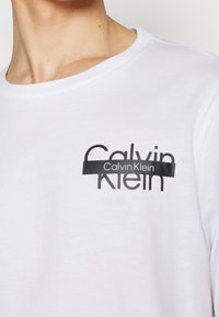 Bílý bavlněný tričko s kulatým výstřihem, s černým grafickým logem "Calvin Klein" na levé části hrudníku. Hladký povrch.