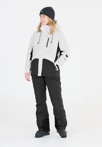 Skijacke in Weiß und Schwarz mit einer Reißverschlusstasche auf der Brust, verstellbarer Kapuze und wasserdichtem Design. Dazu eine schwarze Skihose und dunkle Stiefel.