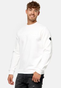 Witte sweatshirt met een ronde halslijn en lange mouwen. Heeft een reliëftekst "INDICODE JEANS" op de voorkant en een zwart logo patch op de mouw.