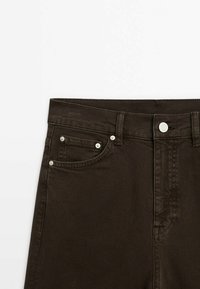 Pantalon en denim brun foncé avec bouton à l'avant et rivets métalliques, montrant la poche avant droite et les passants de ceinture.