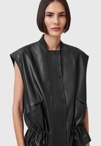 Black sleeveless leather vest with a textured ribbed front, snap buttons, and elasticized waist. Features side pockets and a smooth finish.