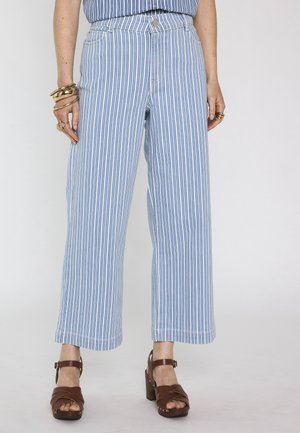 Donna che indossa pantaloni a gamba larga a righe verticali blu e bianche, abbinati a sandali marroni con tacco e braccialetti dorati a una mano.